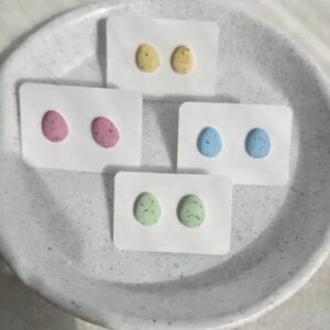 Four pairs of handmade Easter egg stud earrings in pastel yellow, pink, blue and green polymer clay, displayed on white cards on a speckled ceramic plate.