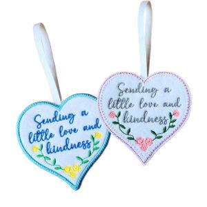 Two heart-shaped felt hanging decorations shown side by side, each with a ribbon loop. Both hearts feature an embroidered message reading, ‘Sending a little love and kindness’, with small floral details.