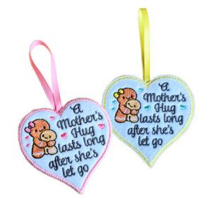 Felt heart hanging decorations with embroidered message 'A Mother’s Hug Lasts Long After She’s Let Go' and a mum and child gingerbread illustration, shown in two colour variations with ribbon loops for hanging.