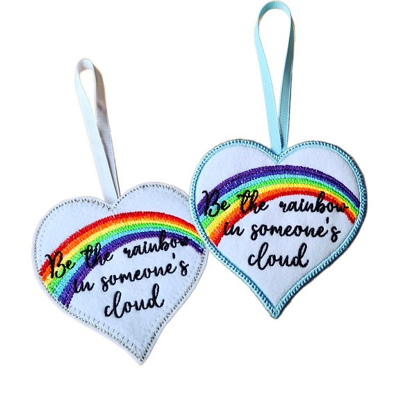Two heart-shaped felt hanging decorations shown side by side, each with a ribbon loop. Both hearts feature an embroidered rainbow and the message ‘Be a rainbow in someone’s cloud’.