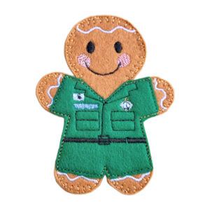 A handmade felt paramedic gingerbread character featuring embroidered black eyes, a smiling mouth, pink cheeks, and decorative icing-style stitching. The character wears a green ambulance uniform with stitched badges, pockets, and a belt detail. Displayed on a clean white background.