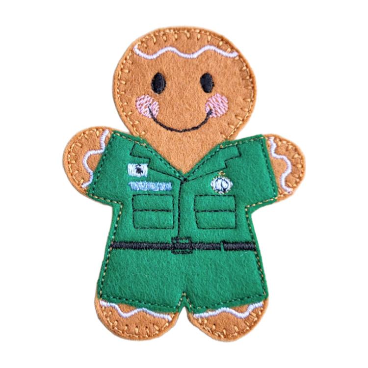 A handmade felt paramedic gingerbread character featuring embroidered black eyes, a smiling mouth, pink cheeks, and decorative icing-style stitching. The character wears a green ambulance uniform with stitched badges, pockets, and a belt detail. Displayed on a clean white background.