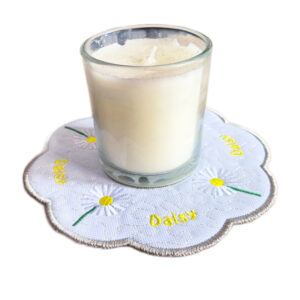 Embroidered daisy candle mat with scalloped edge, featuring white daisies with yellow centres on a white background, holding a glass candle on top.