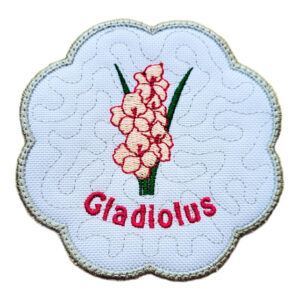 Round scalloped candle coaster embroidered with pink gladiolus flowers and green leaves, featuring a subtle textured background, a soft green stitched border, and the word “Gladiolus” embroidered beneath the design.