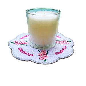 Glass candle placed on a scalloped embroidered gladiolus candle mat, featuring pink gladiolus flowers with green stems, a subtle textured background, and a soft green stitched border with “Gladiolus” embroidered around the design.