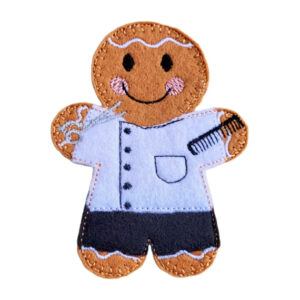 Felt gingerbread barber character wearing a white tunic and black trousers, with embroidered details including a comb and silver scissors, smiling with pink cheeks and decorative icing stitching.