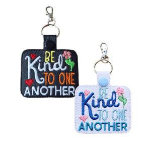 Two embroidered keyrings featuring the phrase “Be Kind to One Another” with colourful hearts and flowers. One keyring is black with yellow, orange, red, green, and white stitching, and the other is white with shades of blue, pink, and green stitching. Both have a snap button, metal ring, and silver lobster clasp for easy attachment.