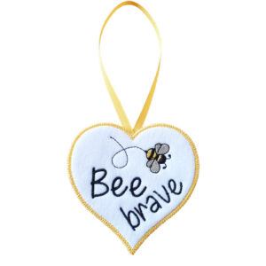 White felt heart-shaped hanging decoration with the words “Bee brave” embroidered in black, featuring a small embroidered bee above the text. The heart has a yellow stitched border and hangs from a matching yellow ribbon loop against a white background.