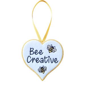 White felt heart-shaped hanging decoration with the words “Bee Creative” embroidered in black, featuring two small embroidered bees. The heart has a yellow stitched border and hangs from a matching yellow ribbon loop against a white background.
