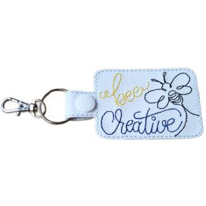 A white rectangular keyring with a metal clasp and ring, embroidered with the words “Bee Creative” in yellow and blue thread. The design includes a simple line drawing of a bee, symbolising creativity and positivity.