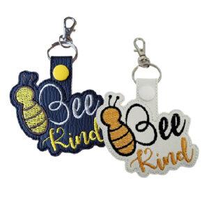 Two embroidered keyrings featuring the phrase “Bee Kind” with a bee design. One keyring is black with yellow and silver stitching and a yellow snap button, while the other is white with orange and black stitching and a white snap button. Both have metal rings and swivel lobster clasps for easy attachment.