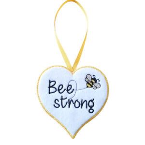 Soft felt heart-shaped hanging decoration with the words “Bee Strong” embroidered in yellow, featuring a small embroidered bee and a yellow stitched border. The heart hangs from a matching yellow ribbon loop against a white background.