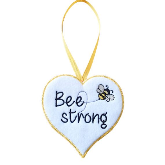 Soft felt heart-shaped hanging decoration with the words “Bee Strong” embroidered in yellow, featuring a small embroidered bee and a yellow stitched border. The heart hangs from a matching yellow ribbon loop against a white background.