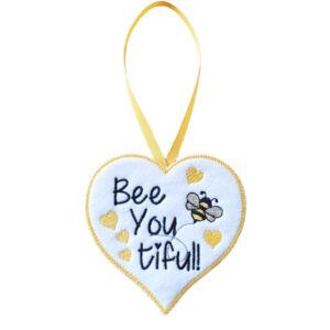 White felt heart-shaped hanging decoration with the words “Bee You tiful!” embroidered in black, featuring a small embroidered bee and several yellow hearts. The heart has a yellow stitched border and hangs from a matching yellow ribbon loop against a white background.