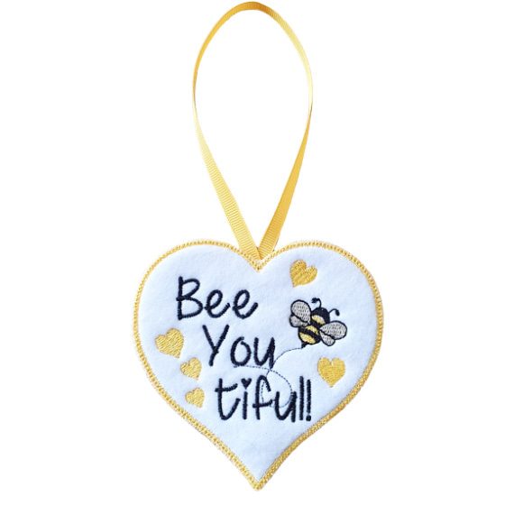 White felt heart-shaped hanging decoration with the words “Bee You tiful!” embroidered in black, featuring a small embroidered bee and several yellow hearts. The heart has a yellow stitched border and hangs from a matching yellow ribbon loop against a white background.