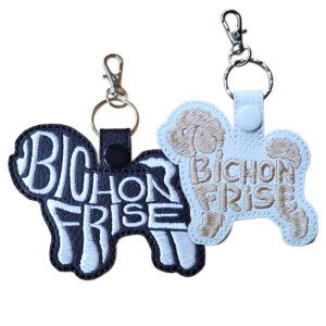Two embroidered Bichon Frise word art keyrings made from faux leather, showing the Bichon Frise dog silhouette with the words “Bichon Frise” stitched into the body. One keyring is black with light stitching, and the other is white with soft neutral stitching, both finished with a snap tab, metal split ring, and lobster clasp.
