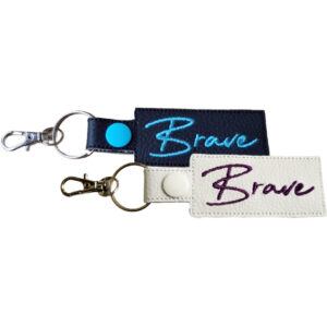 Two rectangular keyrings with the word "Brave" embroidered in cursive. One keyring is black with turquoise embroidery and a silver clasp, and the other is white with dark purple embroidery and a silver clasp.
