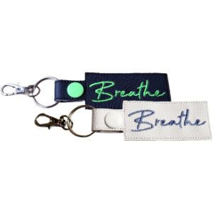 Two rectangular keyrings with the word "Breathe" embroidered in cursive. One keyring is black with lime green embroidery, while the other is white with blue embroidery. Both keyrings have a metal clasp attached to a split ring.