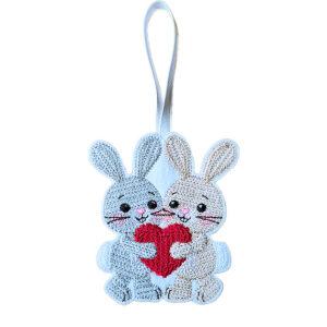 Two felt bunnies hugging and holding a red heart, machine embroidered with a knit stitch pattern and hanging from a ribbon on a white background