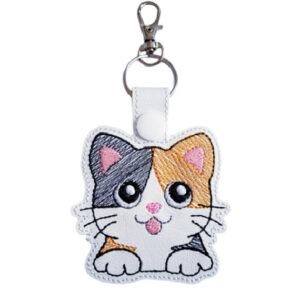 A cute embroidered keyring shaped like a cartoon calico cat with big shiny eyes, a pink nose, and its tongue sticking out. The cat has orange, black, and white patches, and the keyring includes a silver clasp and ring at the top for attaching to keys or bags.