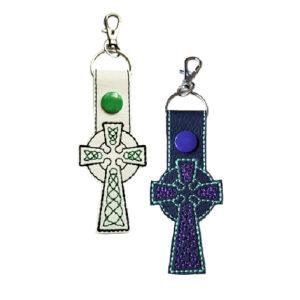Two embroidered Celtic Cross keyrings made from faux leather, one white with green stitching and one black with purple stitching, each featuring a snap button and metal clasp for attaching to keys or bags.