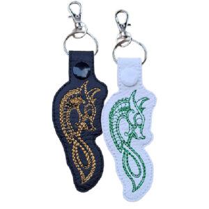 Two Celtic Dragon keyrings crafted from faux leather, one black with gold embroidery and one white with green embroidery, each featuring intricate Celtic knotwork and a snap fastener, symbolising power, protection, and ancient wisdom.