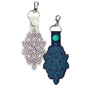 Two faux leather keyrings with intricate embroidered Celtic knot designs, one in white with purple stitching and one in black with green stitching, each with a metal clasp and snap button fastening.