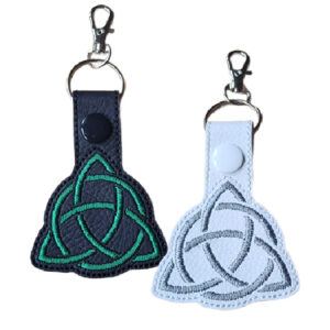 Two embroidered Celtic Triquetra keyrings made from faux leather, one black with green stitching and one white with grey stitching, each featuring a snap button and metal clasp for attaching to keys or bags.