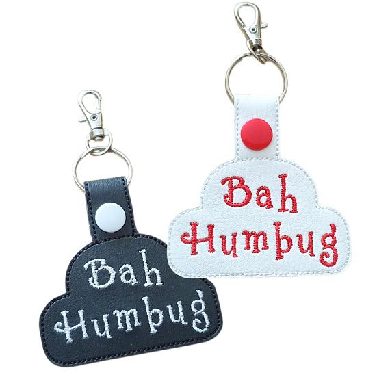Two faux leather Bah Humbug keyrings in cloud shapes, one black with white embroidered text and one white with red embroidered text, each with a metal clasp and snap button fastening.