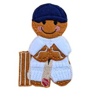 Felt gingerbread cricket player character wearing a navy cap and white cricket kit, holding a bat with pads and a small red cricket ball at the feet, shown standing beside brown cricket stumps.