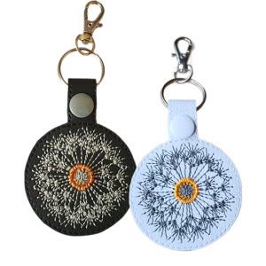 Dandelion clock keyrings shown side by side, one in black and one in white, each featuring a circular dandelion seed head design and attached to a metal keyring ring with a lobster clasp.