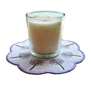 Glass candle placed on a round candle mat with scalloped edges, embroidered with delicate dandelion clock designs and a lilac stitched border.