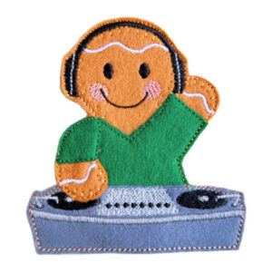 A felt gingerbread character wearing headphones stands behind a DJ deck. The character has a smiling face with rosy cheeks and white stitched icing details. The gingerbread is wearing a green top, and the DJ deck is stitched in shades of grey.