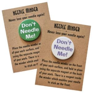Two round magnetic needle minders displayed on kraft backing cards with the heading "Needle Minder – Never lose your needle again!" One minder has a green background with bold white text that reads “Don’t Needle Me!”, while the other has a white background with purple text that reads the same phrase.