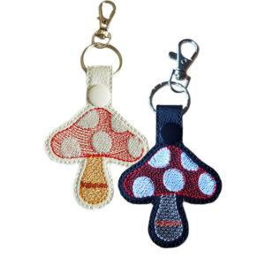 Two embroidered toadstool keyrings made from faux leather, one white with red and beige stitching and one black with red and white stitching, each attached to a metal keyring and lobster clasp.