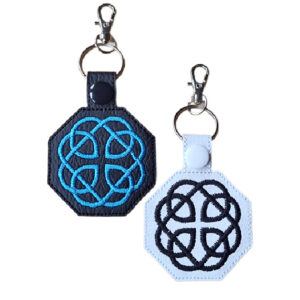Two embroidered Fatherhood Celtic Knot keyrings made from faux leather, one black with blue stitching and one white with black stitching, each featuring a snap button and metal clasp for attaching to keys or bags.