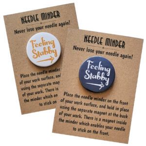 Two round needle minders, one with a white background and orange text, and one with a dark grey background and white text, both reading “Feeling Stabby” with an arrow design, displayed on kraft backing cards with usage instructions.