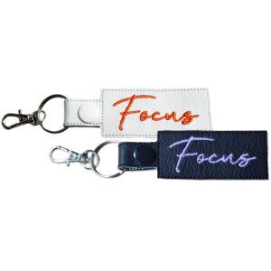 Two rectangular keyrings with the word "Focus" embroidered in cursive. One keyring is white with orange embroidery and a silver clasp, and the other is black with purple embroidery and a silver clasp.