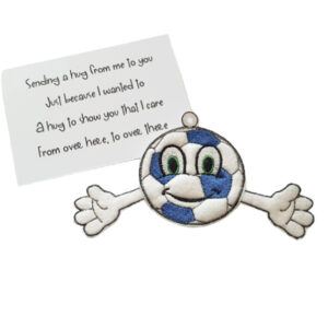 Embroidered football pocket hug charm with a smiling face, green eyes, and outstretched arms, made from soft felt in white and blue. The charm is displayed next to a small keepsake poem card that reads: ‘Sending a hug from me to you, just because I wanted to. A hug to show you that I care, from over here, to over there’.