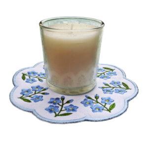 Glass candle displayed on embroidered Forget Me Not decorative candle mat with scalloped edge, blue flowers symbolising love and remembrance, and light blue border.