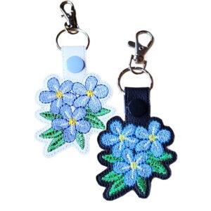 Forget-me-not flower keyrings shown side by side, one in white and one in black, each featuring a blue forget-me-not flower design and attached to a silver keyring ring with a lobster clasp.