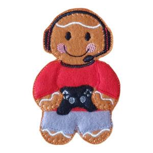 A felt gamer gingerbread character wearing a red top, grey trousers, and a black gaming headset, smiling while holding a small black game controller.