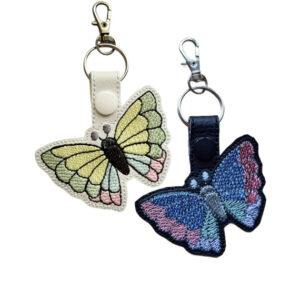 Two embroidered butterfly keyrings made from faux leather, one in white with soft pastel green, yellow, and pink wings, and one in black with blue and pink wings, each finished with a metal keyring clip.