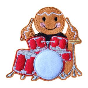 A felt gingerbread character is sitting behind a red drum kit, holding drumsticks in both hands. The gingerbread has a smiling face with rosy cheeks and embroidered icing-style details. The drum kit includes several red drums and a white circular drum head at the front.