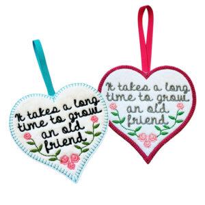 Two heart-shaped felt hanging decorations shown side by side, each with a ribbon loop. Both hearts feature an embroidered friendship message reading, ‘It takes a long time to grow an old friend’, with small decorative flowers.