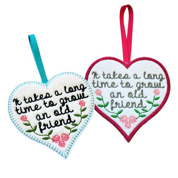 Two heart-shaped felt hanging decorations shown side by side, each with a ribbon loop. Both hearts feature an embroidered friendship message reading, ‘It takes a long time to grow an old friend’, with small decorative flowers.