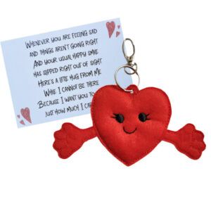 A red felt heart pocket hug charm with a smiling face and outstretched arms, attached to a silver keyring and clasp. Behind it is a keepsake card with a heartfelt poem, sharing comforting words to show care and support.