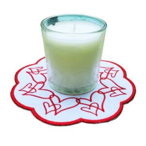 White scalloped decorative table mat with a red embroidered heart wreath design, shown beneath a clear glass candle holder filled with white wax, demonstrating use as a candle mat.