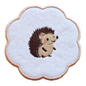 Embroidered candle coaster with a scalloped edge, featuring a single hedgehog with brown spines and a beige body on a white background with a rose gold border.