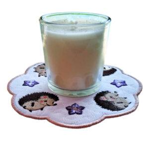 Glass candle placed on an embroidered circular candle mat with a scalloped edge, featuring four hedgehogs and four purple flowers on a white background with a rose gold border.